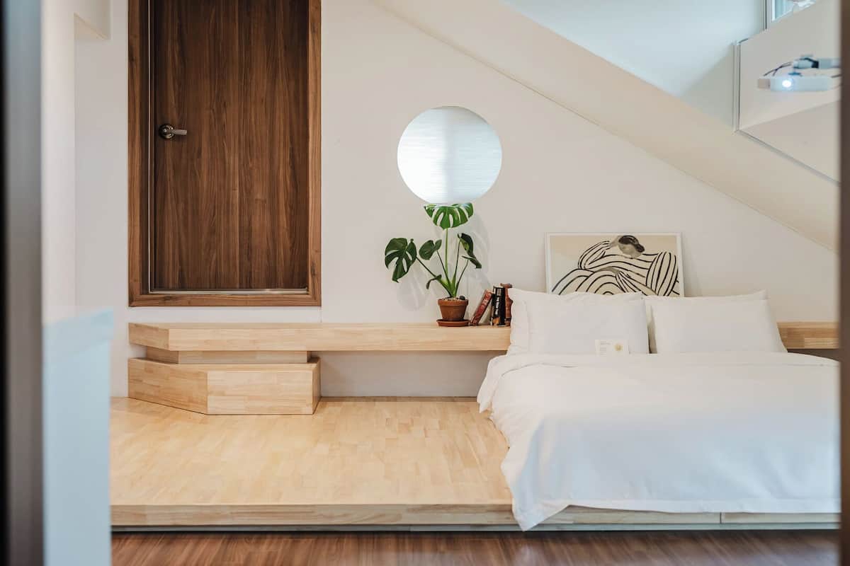 Ein Schlafzimmer mit einem Bett, einer Holztür, einem Topfpflanzen, einem wandmontierten Spiegel und einem gerahmten Kunstwerk.