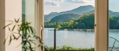 A view from a room with a large window overlooking a lake, surrounded by greenery and mountains.
