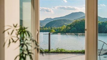 Vista desde una habitación con un ventanal grande que da a un lago, rodeado de vegetación y montañas.