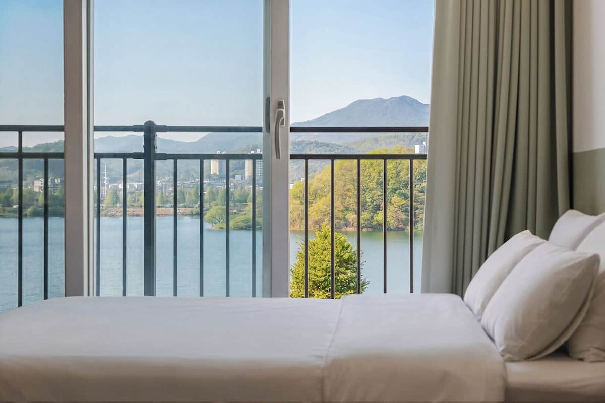 Ein Hotelzimmer mit großem Fenster, einem Bett und Blick auf einen See und Berge.