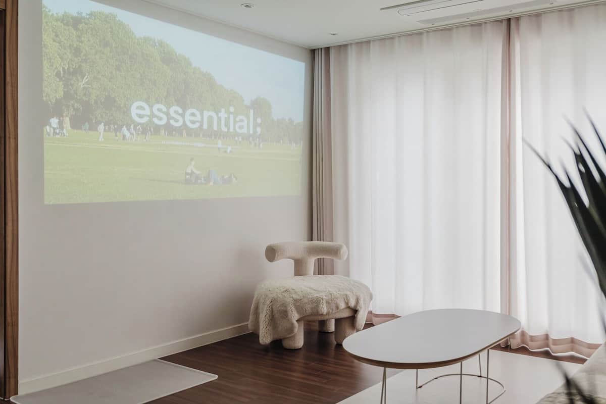 Ein Zimmer mit einer Projektionsleinwand, die 'essential' zeigt; ein bequemer Sessel, ein kleiner Tisch und eine große Pflanze.