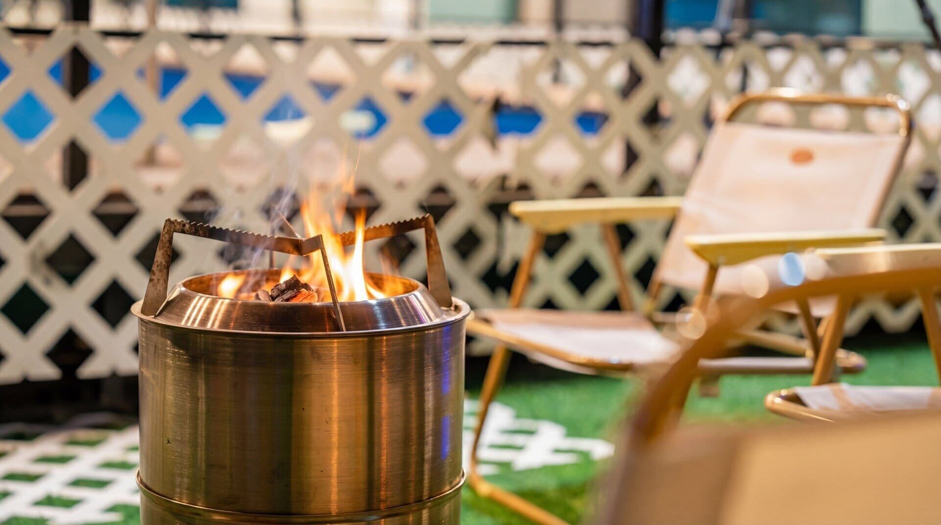 A portable stove with a metal pot on a green surface, a wooden chair, and a lattice fence in the background.