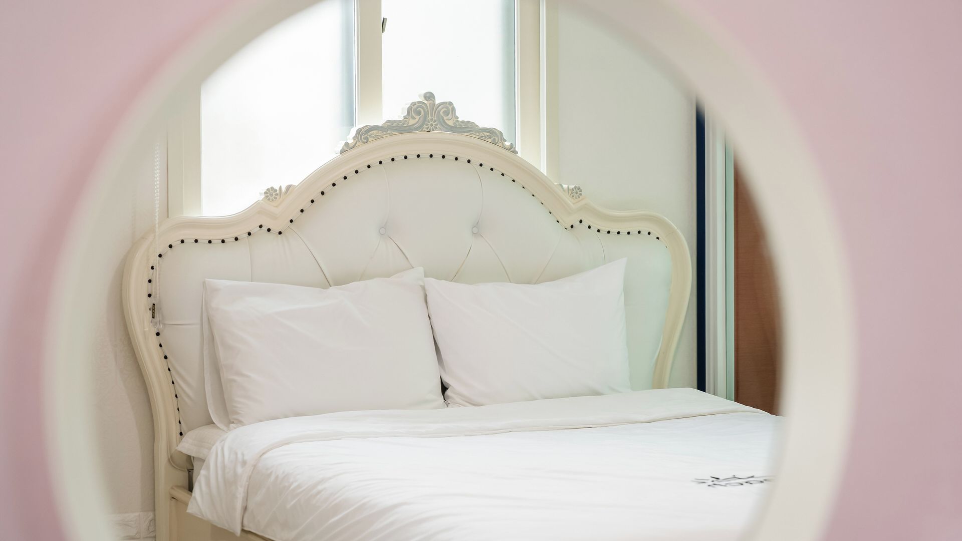 Un lit avec une tête de lit blanche et des oreillers, une fenêtre donnant sur un beau paysage et un mur rose.