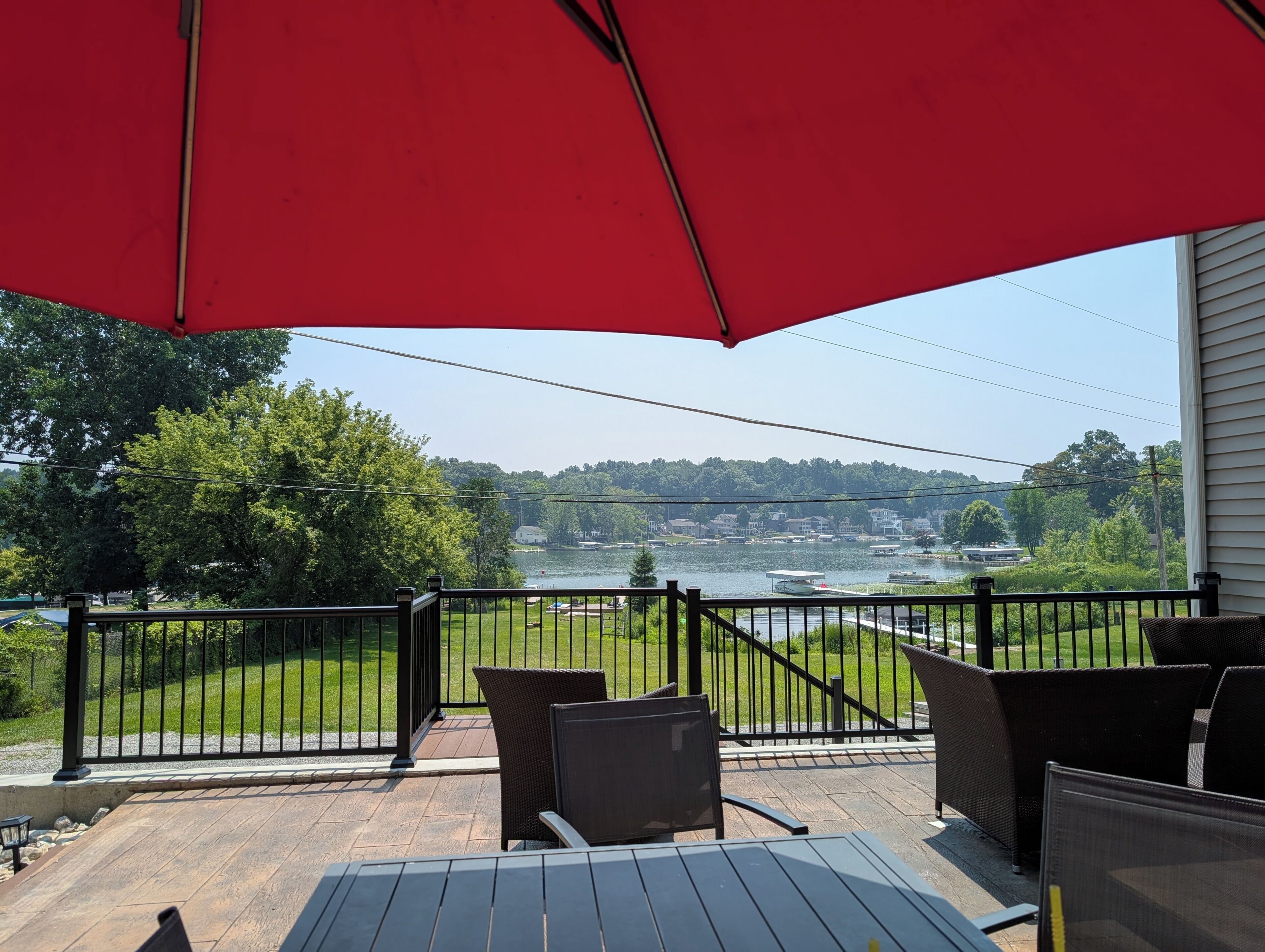 Une terrasse avec des meubles d’extérieur, un parasol rouge et une vue sur un lac, avec des bateaux et des maisons en arrière-plan.