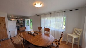 Uma área de refeições com mesa e cadeiras de madeira, cozinha ao fundo e vista para o verde lá fora.