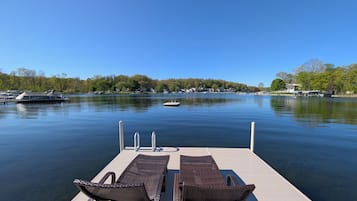 A dock with two wicker lounge chairs facing a calm lake, surrounded by trees and houses.