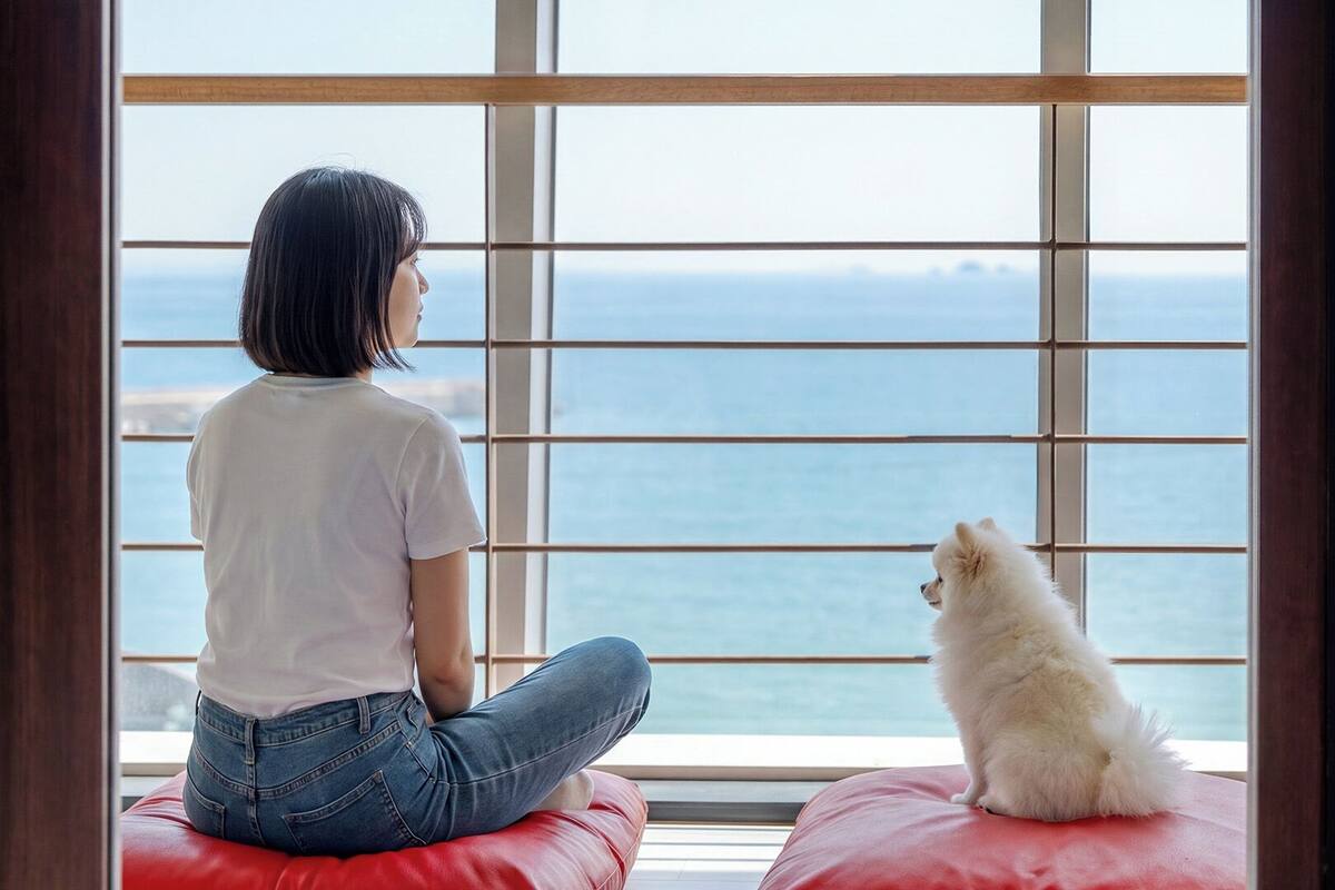 Una persona sentada sobre un cojín rojo junto a una ventana con vista al mar.