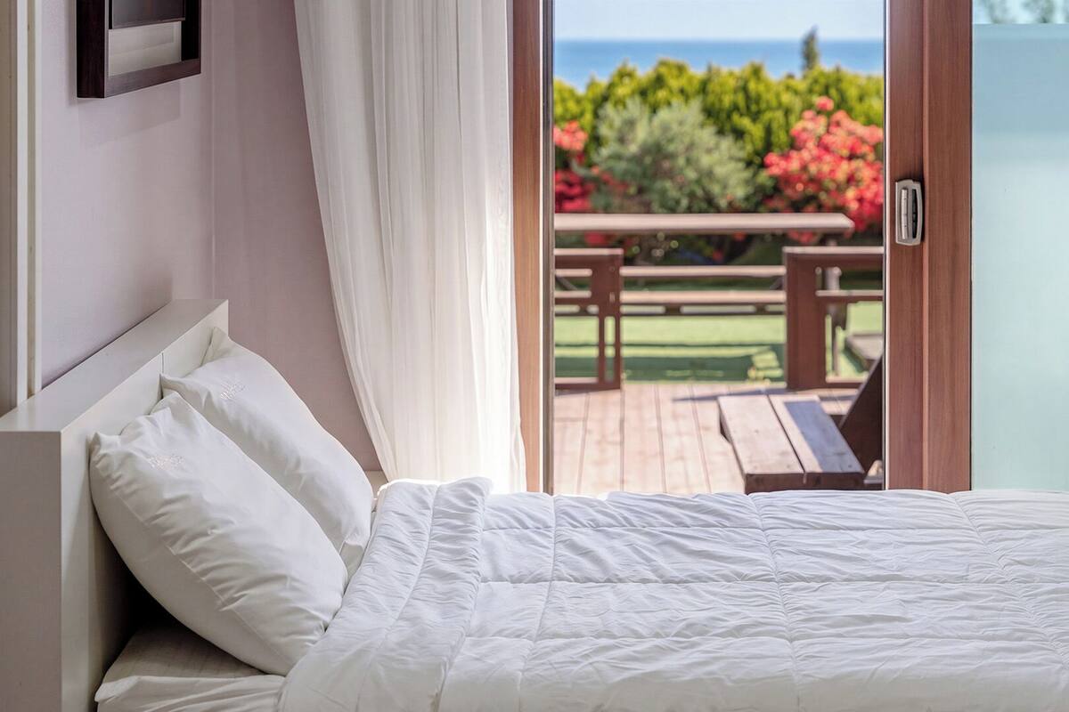 Una cama con ropa blanca, cabecera de madera y vistas a una terraza exterior con mesa y sillas.