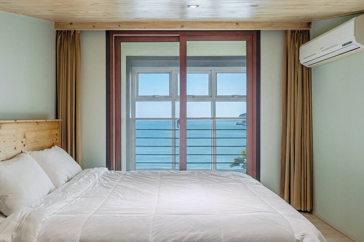 Un dormitorio con cabecera de madera, cama con ropa de cama blanca, un ventanal con vistas al mar y un equipo de aire acondicionado en la pared.