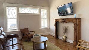 Una sala de estar con un sofá de cuero, una mesa de centro de madera, una silla de mimbre, una chimenea y un televisor montado en la pared.