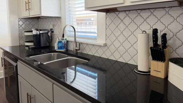 Una cocina con gabinetes blancos, encimera negra, pileta de acero inoxidable, cafetera y bloque para cuchillos.