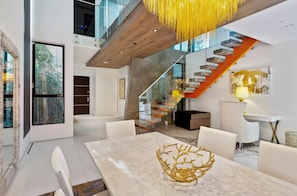 Un interior moderno con una mesa de comedor de mármol, una escalera con barandillas de vidrio y vistas a zonas verdes exteriores.