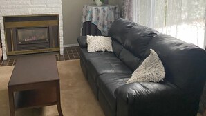 A living room with a fireplace, a black leather sofa, a small table, and a star decoration on the wall. - Family Friendly Cottage close to beach and main strip. (Grand Bend)