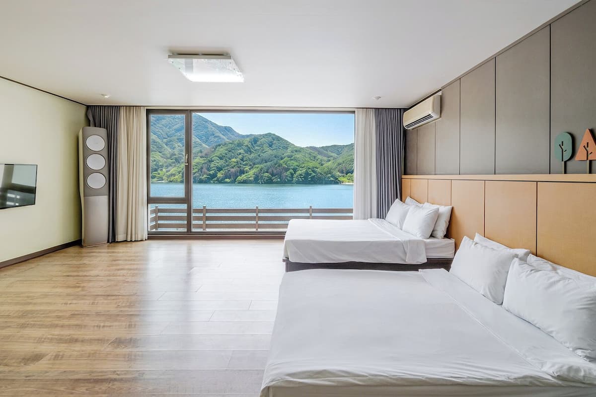 A hotel room with two beds, a TV, a large window with a view of a lake and mountains, and a ceiling fan.