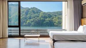 Une chambre d’hôtel avec une grande fenêtre, un lit, une petite table et une vue sur une colline boisée.