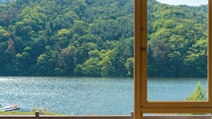 Une vue à travers une fenêtre sur un lac entouré d’une forêt dense, avec un cadre en bois visible au premier plan.