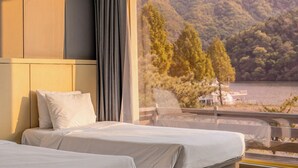 Une chambre d’hôtel avec un lit, une fenêtre donnant sur un lac et des arbres, et un balcon avec une balustrade.