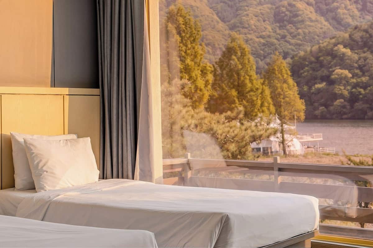 A hotel room with a bed, a window with a view of a lake and trees, and a balcony with a railing.
