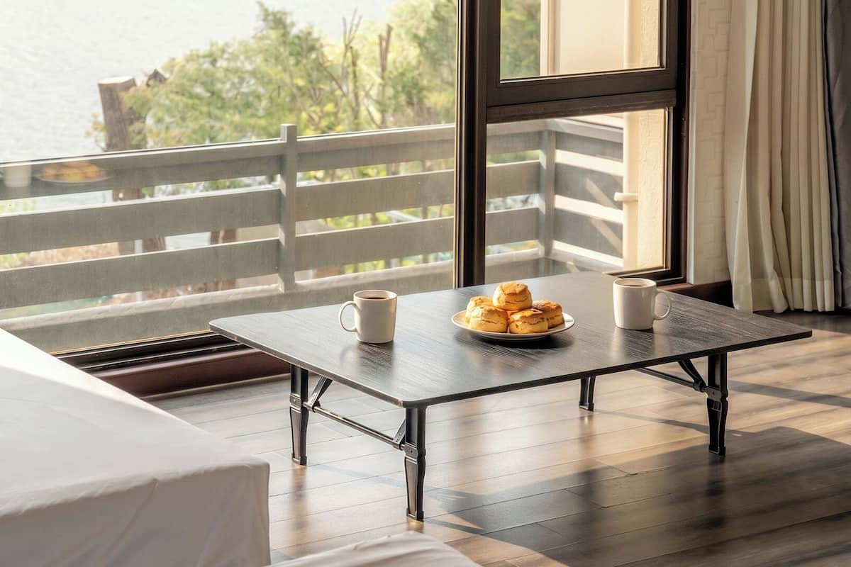 A room with a bed, a table with a plate of pastries and two cups, and a view of the outdoors.