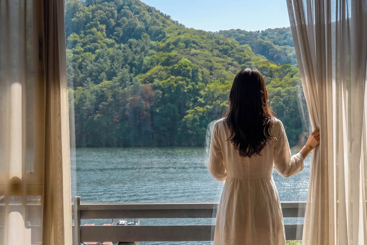 A person standing by a window with curtains, overlooking a body of water and a forested hillside.
