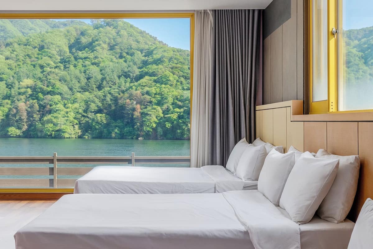 A hotel room with two beds, a large window with a view of a forested lake.