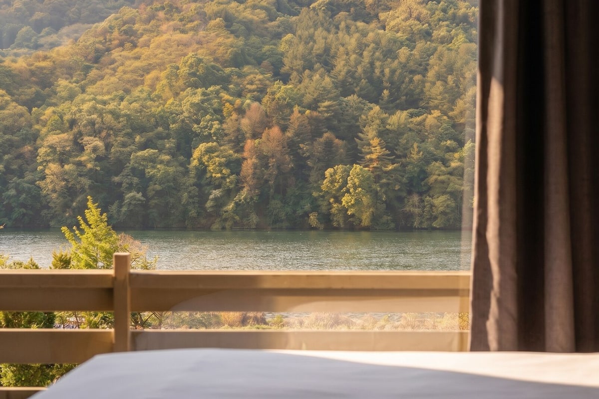 A room with a bed, a window overlooking a forested area and a body of water.