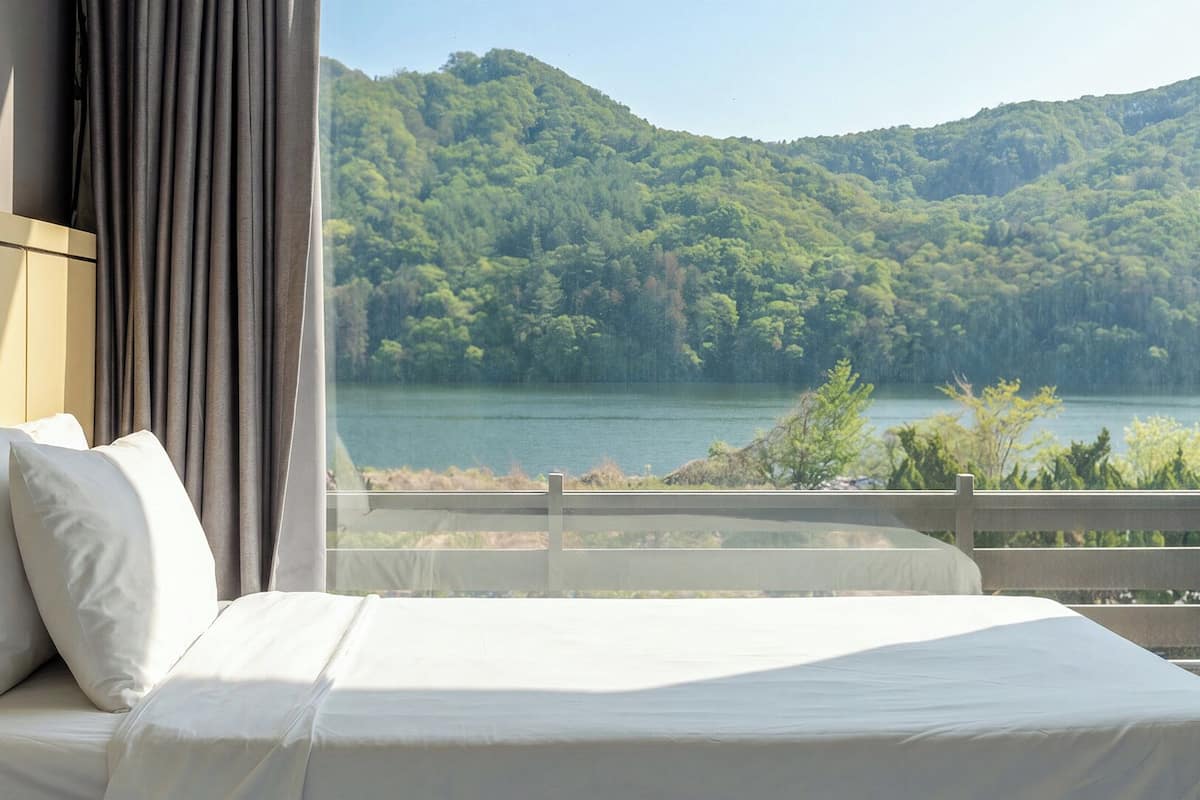 A hotel room with a bed, white bedding, a view of a lake and forested hills.