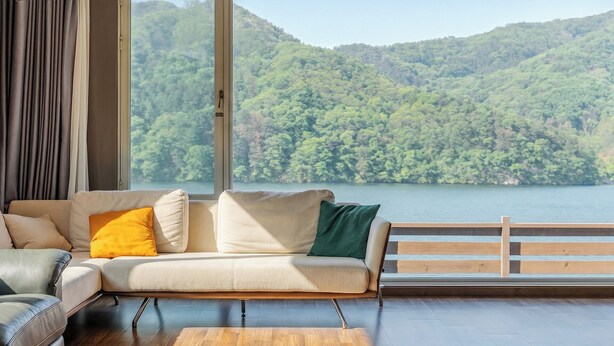 Un salon avec un canapé, un plancher en bois et une grande fenêtre donnant sur un lac et des collines boisées.