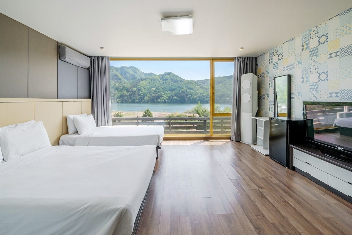 A hotel room with two beds, a TV, and a view of a lake and mountains.