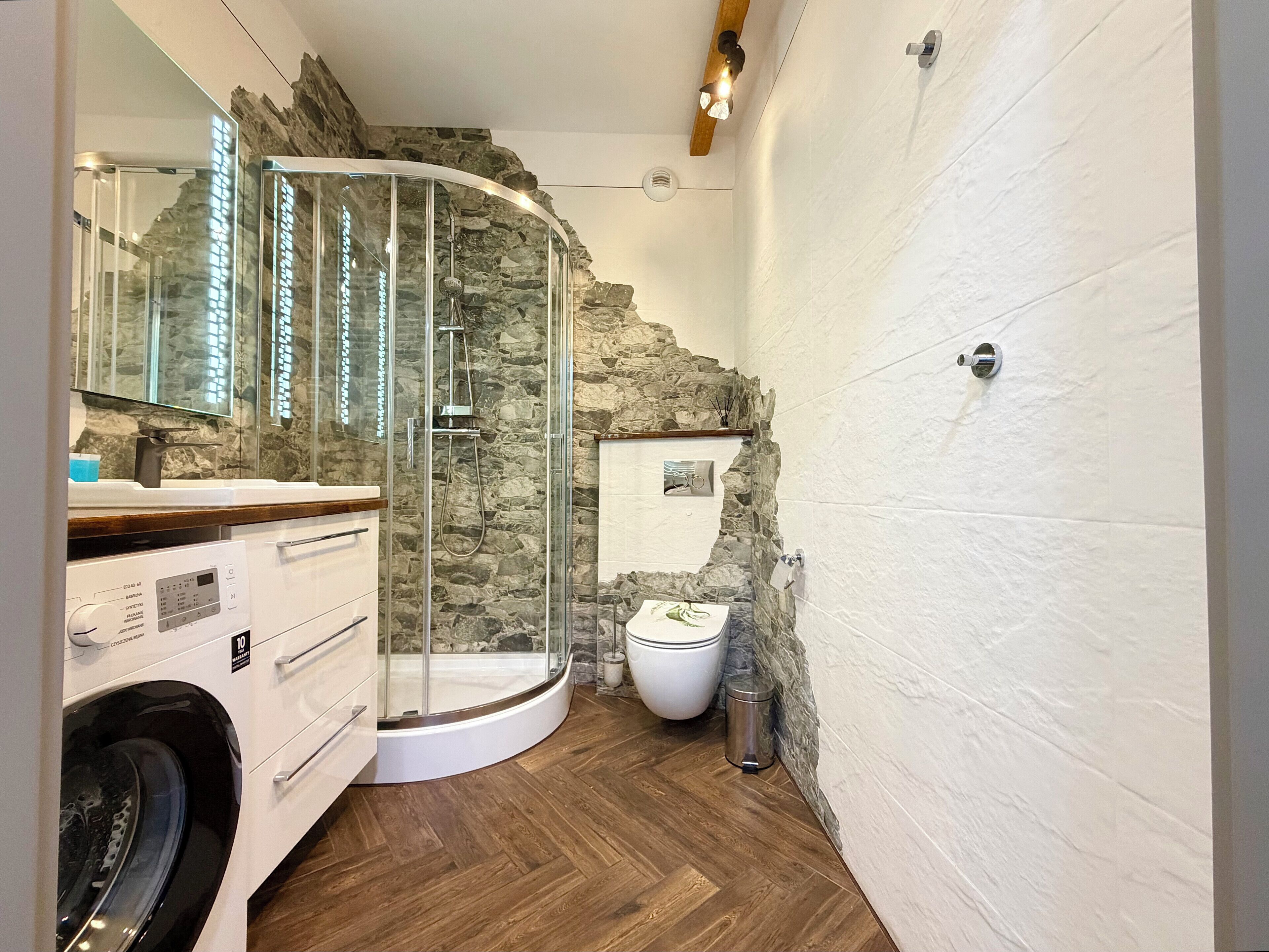 A modern bathroom with a stone wall, a glass shower enclosure, a washing machine, and a toilet.