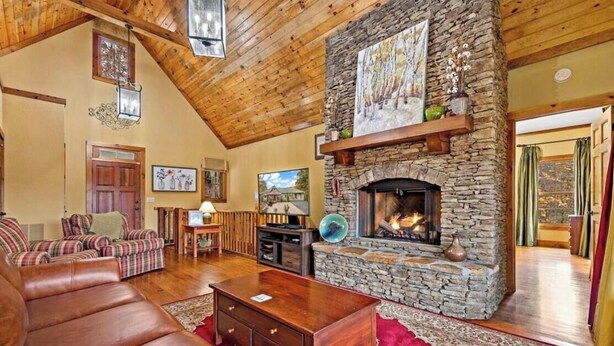 A cozy living room with a stone fireplace, wooden ceiling, and plaid furniture. - Peaceful perfection! 4-bedroom mountain lake house on Lake Adger. Near Lake Lure (Mill Spring)