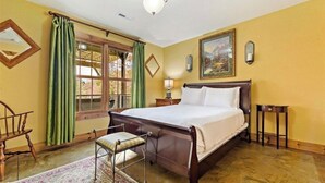 A bedroom with a wooden bed, a chair, a nightstand, a lamp, a painting, and a window with curtains. - Peaceful perfection! 4-bedroom mountain lake house on Lake Adger. Near Lake Lure (Mill Spring)