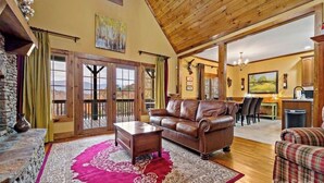 A spacious living room with a leather sofa, a coffee table, and a red patterned rug. There is a stone fireplace, wooden ceiling beams, and a view of the outdoors through large sliding doors. - Peaceful perfection! 4-bedroom mountain lake house on Lake Adger. Near Lake Lure (Mill Spring)