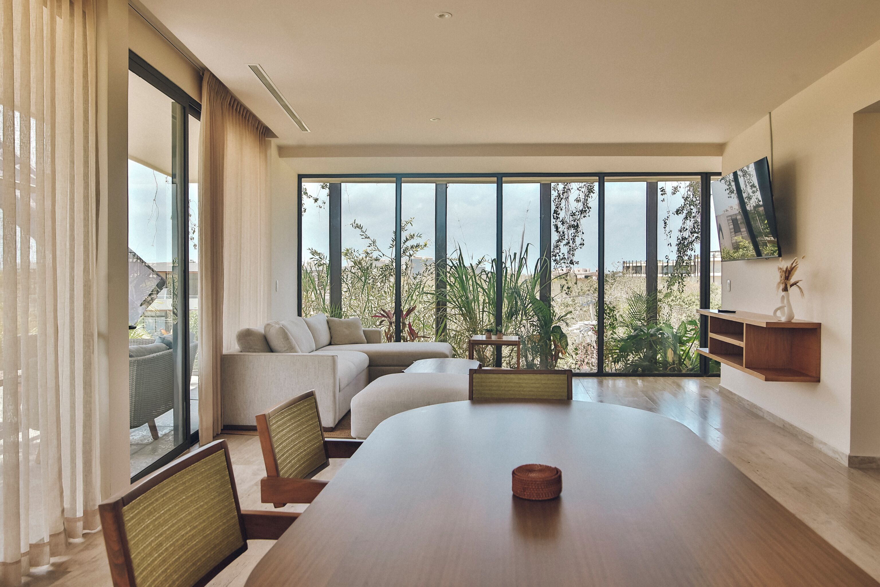 Un salón moderno con una gran mesa de comedor, asientos cómodos, un televisor montado en la pared y vistas a zonas verdes y edificios a través de amplios ventanales.