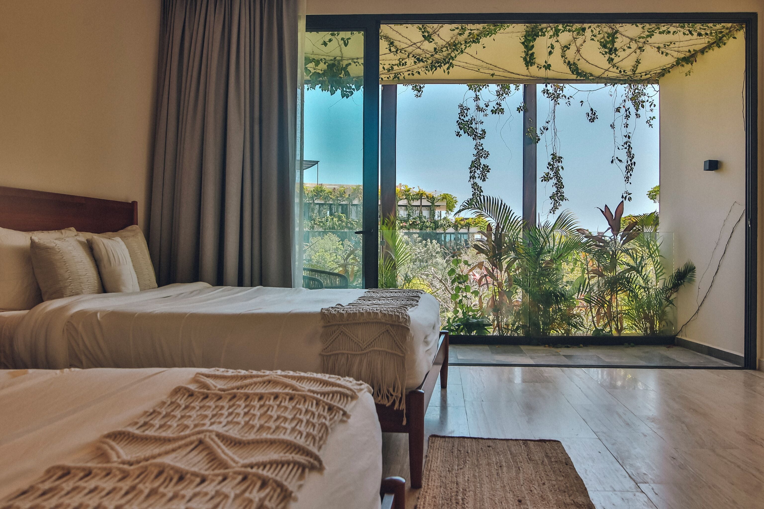 Un dormitorio con dos camas, un amplio ventanal corredera que da a un balcón con plantas y vistas a edificios.