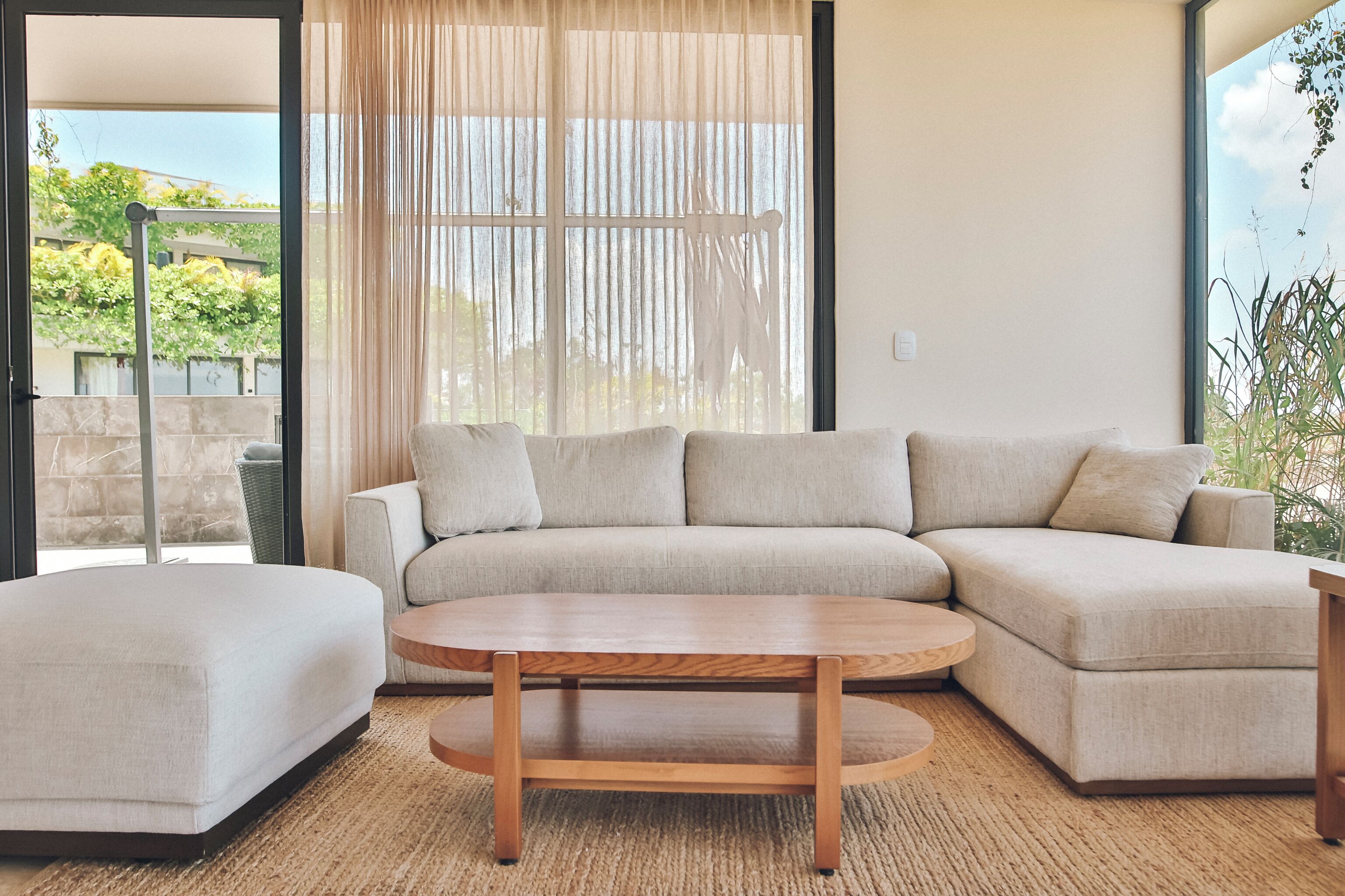 A modern living room with a sectional sofa, a round wooden coffee table, and a large window with sheer curtains.