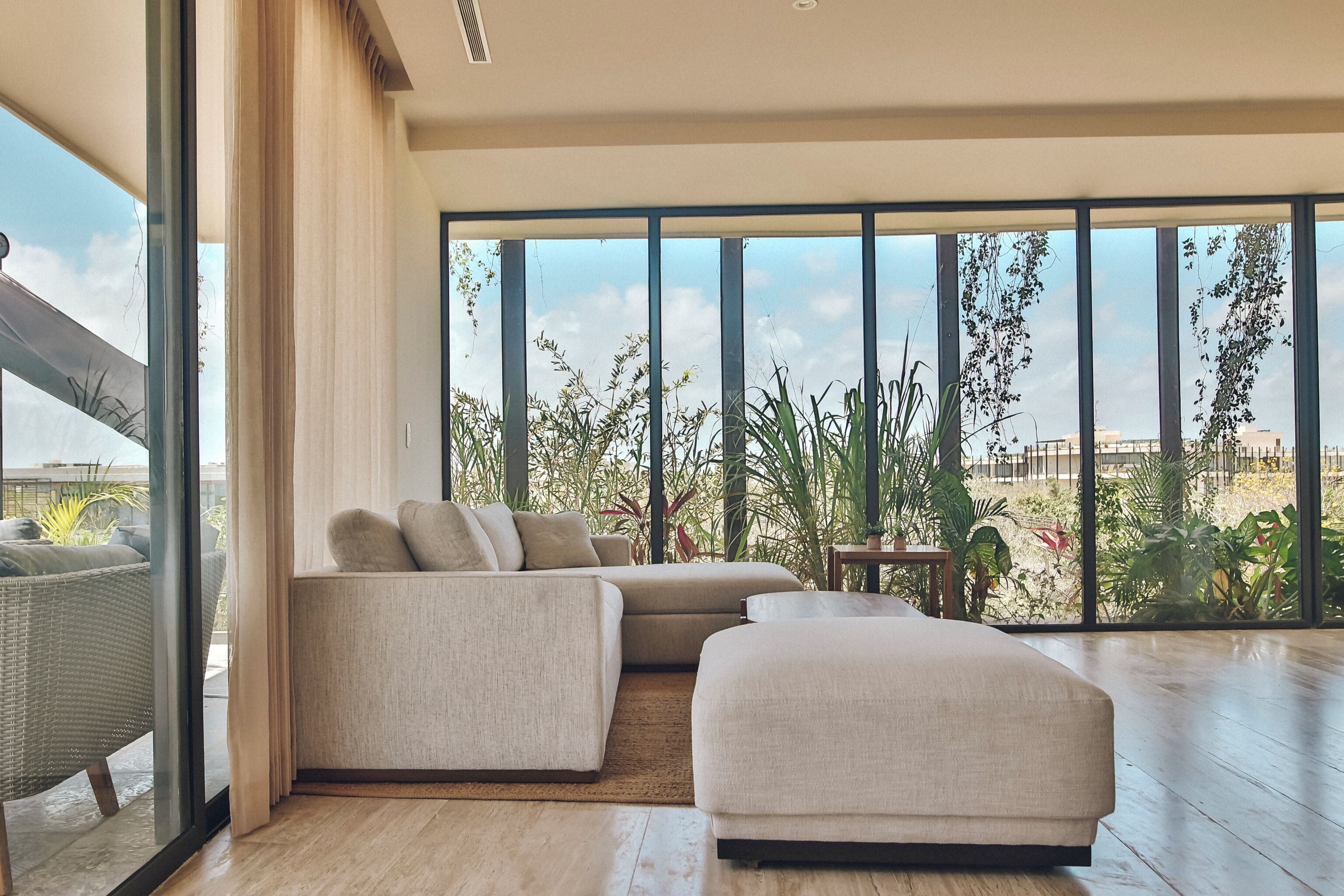Una sala de estar moderna con un sofá seccional, un pequeño reposapiés y vistas a un exuberante jardín a través de grandes ventanales.