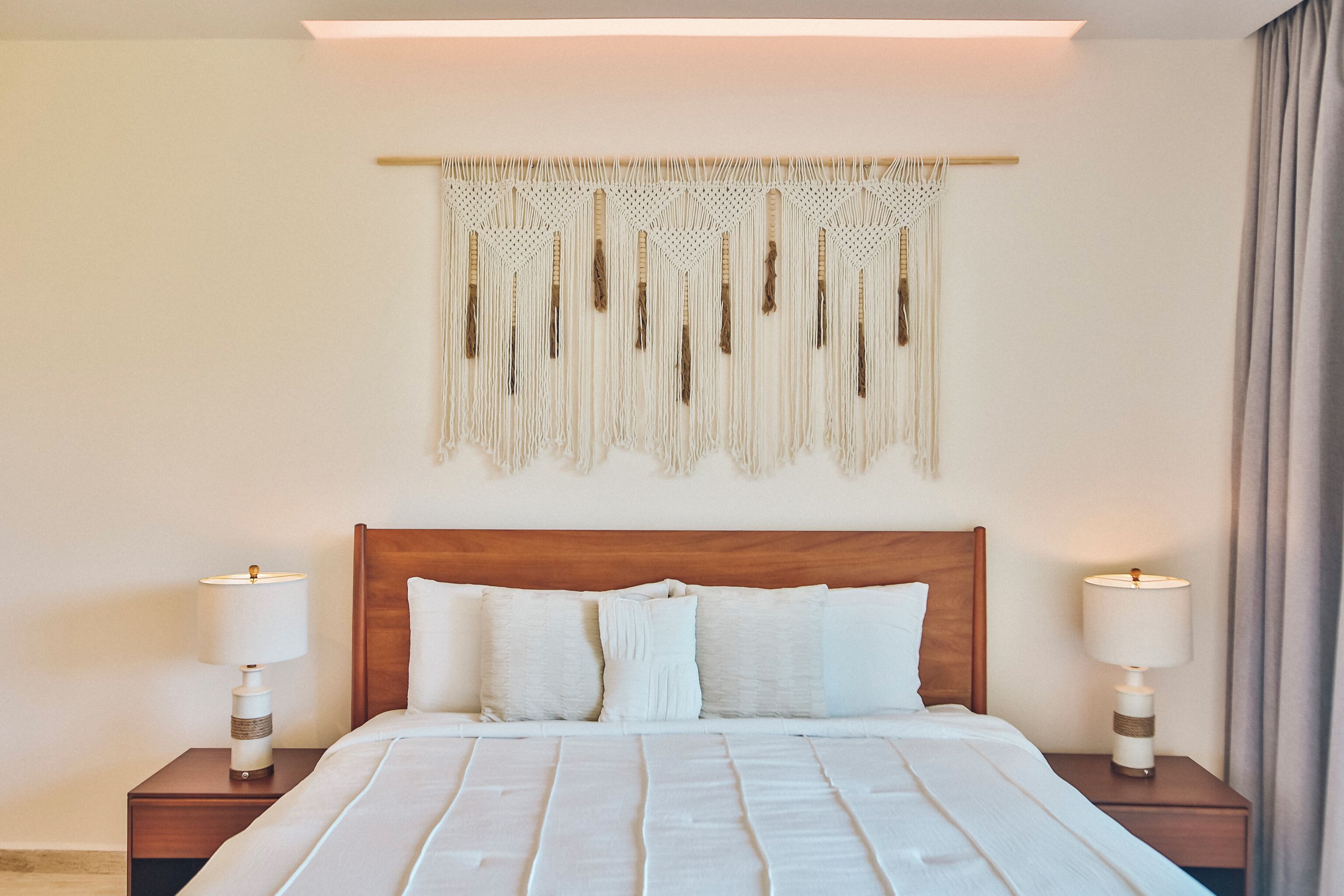 A hotel room with a wooden headboard, a bed with white bedding, two bedside tables with lamps, and a macrame wall hanging.
