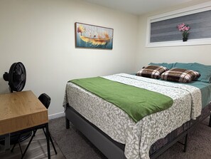 Ein Schlafzimmer mit einem Bett, einem Schreibtisch, einem Stuhl, einem Standventilator und zwei Wandbildern.
