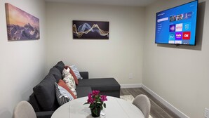 Una sala de estar con un sofá gris, una mesa redonda con planta en maceta y un televisor de pantalla plana que muestra servicios de streaming.
