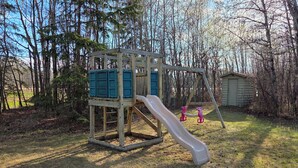 Une structure de jeu en bois, dotée d’un toboggan et d’une zone d’escalade, entourée d’arbres et d’un petit abri de jardin.