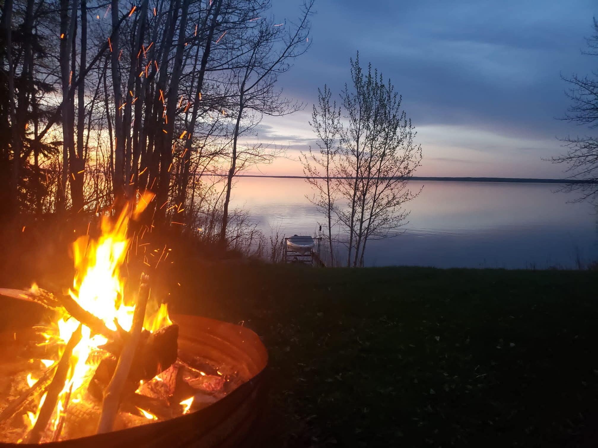 A campfire in a metal bowl, a calm lake, and leafless trees.