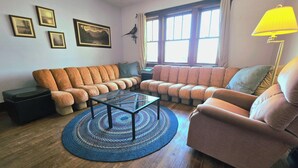Un salon comprenant un canapé d’angle, une table basse en verre, un tapis à motifs et une fenêtre avec des rideaux.