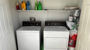 Een wasserette met twee witte voorladers, een plank met verschillende schoonmaakproducten en een opbergruimte met zakken en blikken.