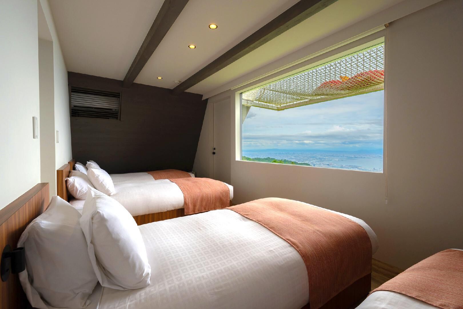 A hotel room with two beds, a large window with a view, and a ceiling with exposed beams and recessed lighting.