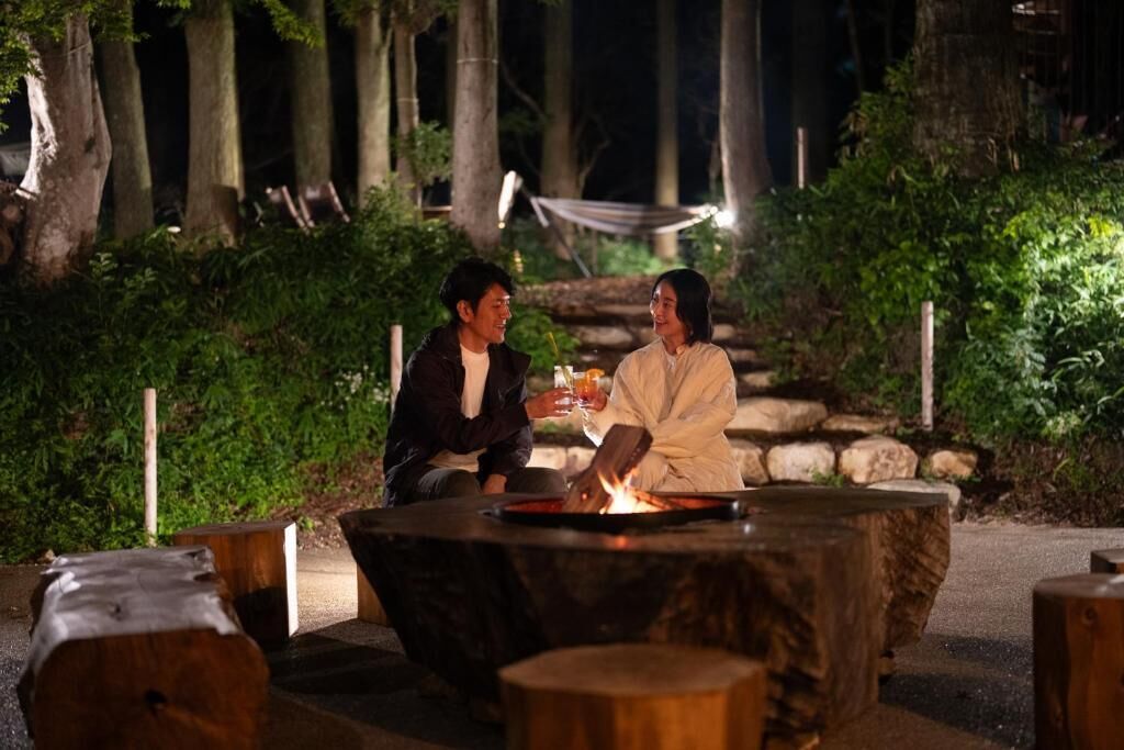 Two people sitting at an outdoor table with a fire, surrounded by greenery and trees.