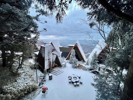 A snow-covered outdoor area with unique triangular buildings, a central circular seating arrangement, and a person in red attire.. Nature Live Rokko