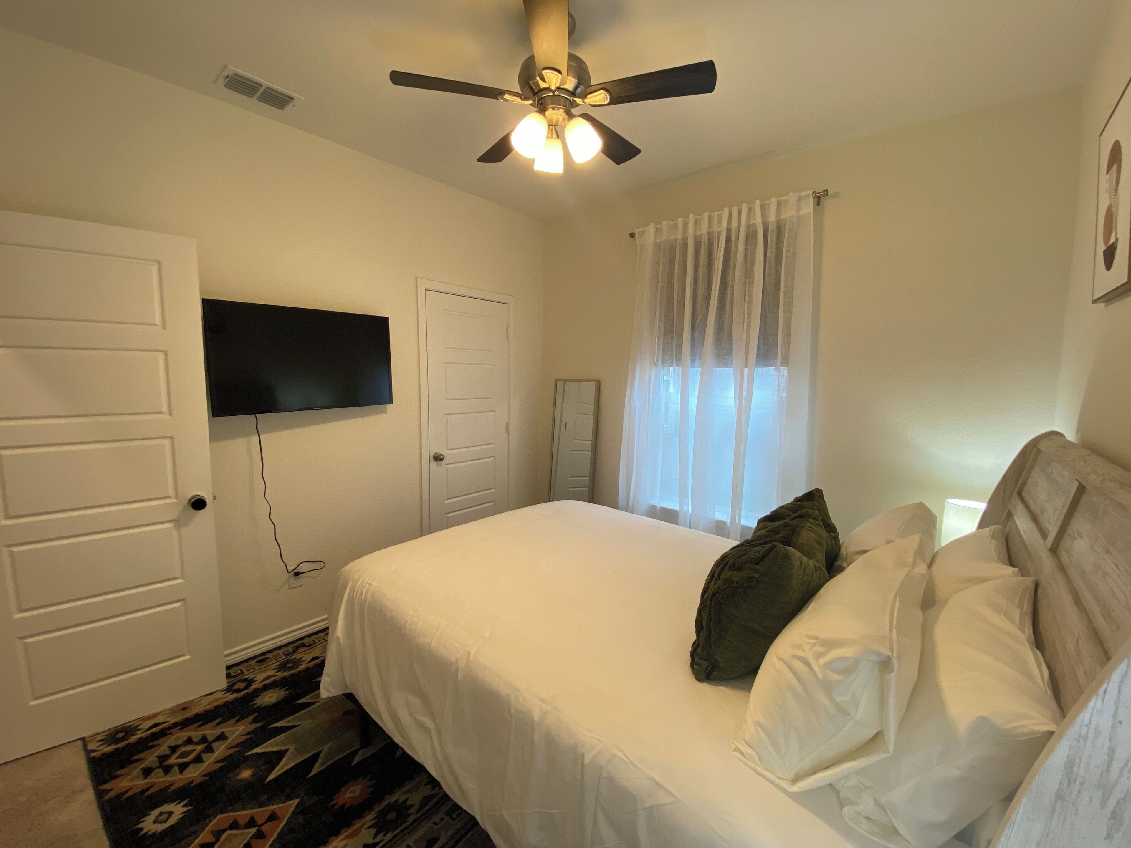 Quarto com cama, ventoinha no teto, televisão na parede e janela com cortinas.
