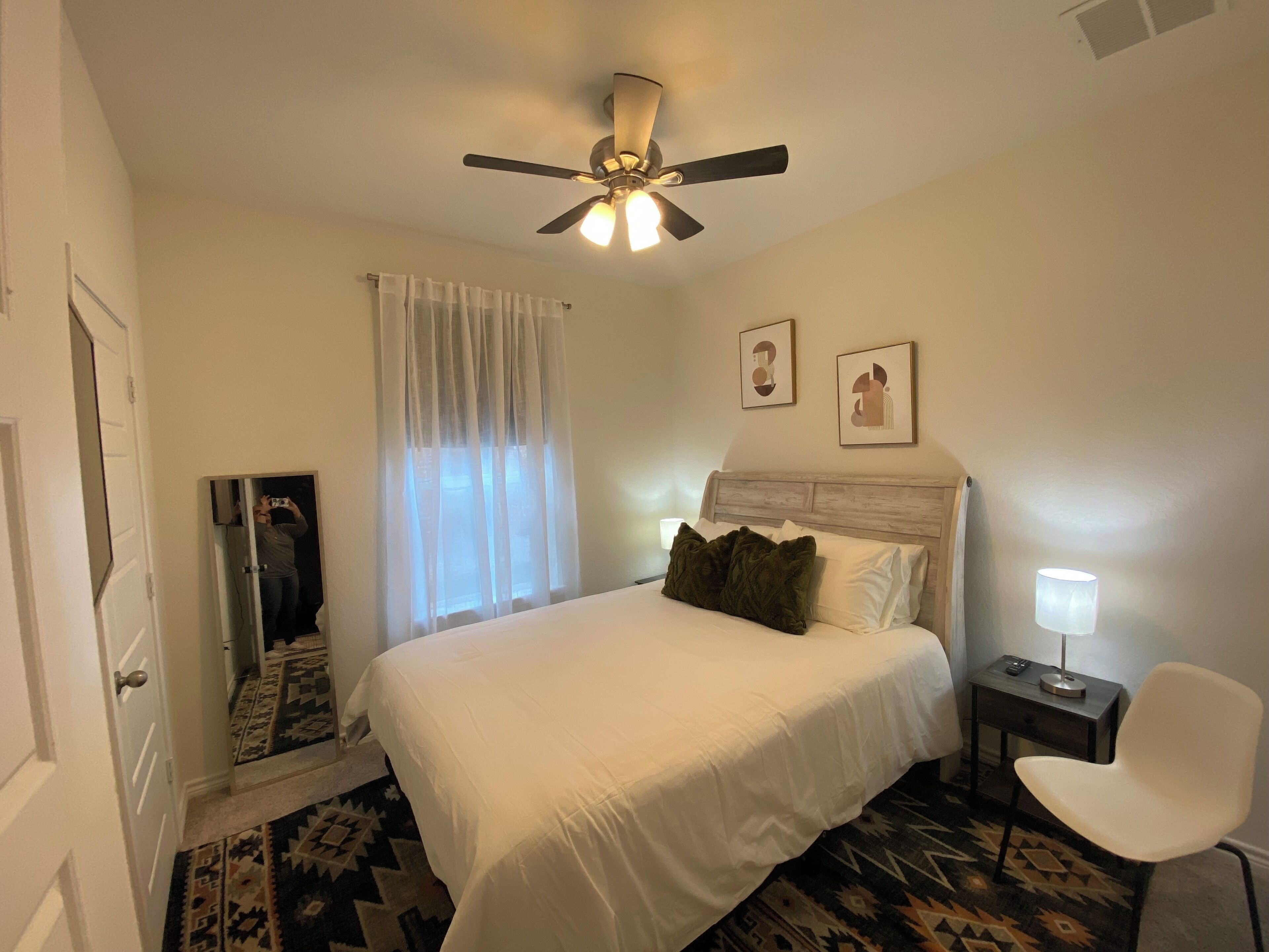 A hotel room with a bed, a ceiling fan, a nightstand, a chair, a window with sheer curtains, and a rug.