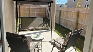 Een overdekt terras met twee stoelen en een kleine tafel, een houten schutting en huizen op de achtergrond.